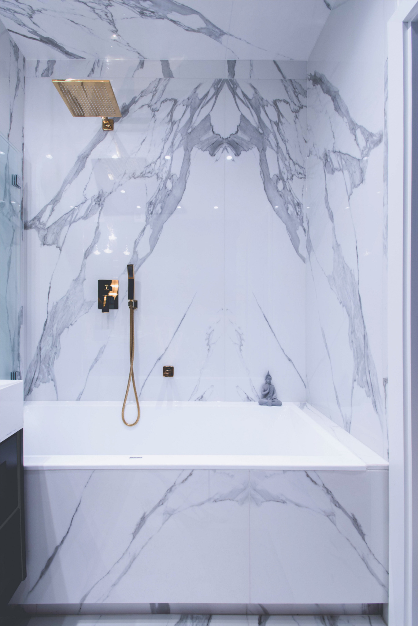 Nowoczesna łazienka wyłożona białym marmurem z szarymi żyłami, złota deszczownica i bateria, minimalistyczna wanna z figurką Buddy.