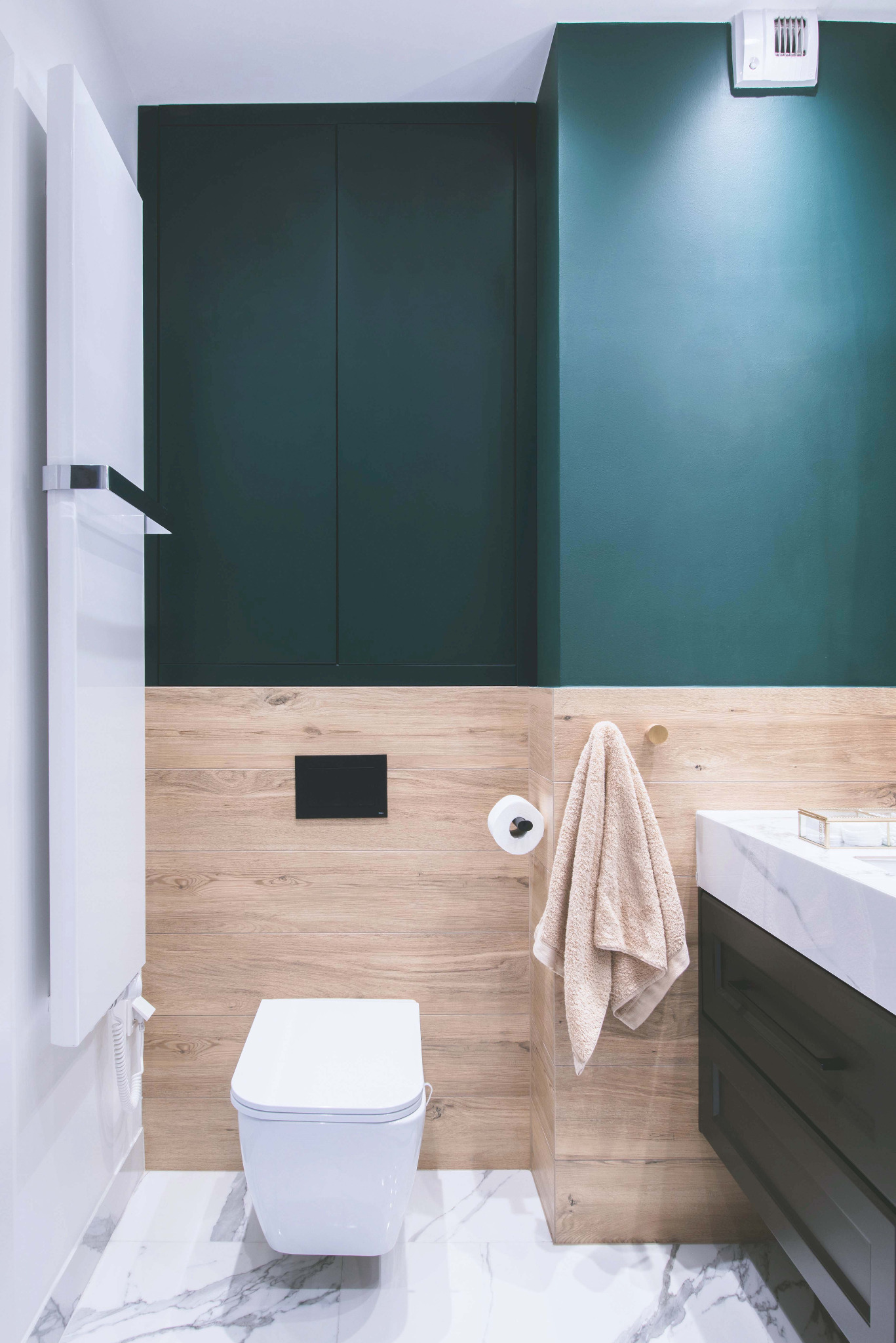 Nowoczesna łazienka z białym kompaktem WC, drewnopodobnymi płytkami ściennymi, marmurową posadzką i ciemnozieloną ścianą z szafką w zabudowie.