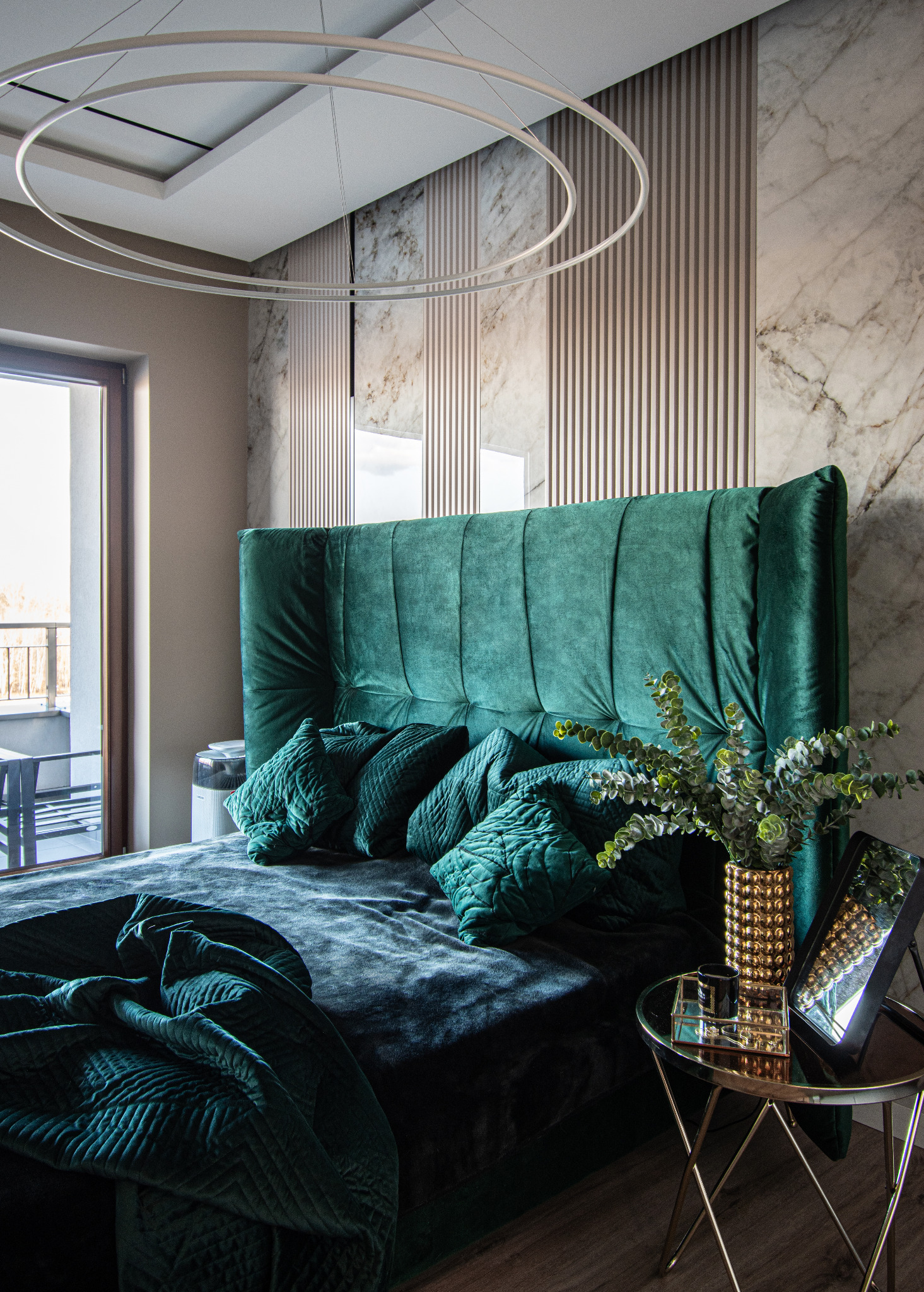 Elegancka sypialnia z wysokim, szmaragdowym zagłówkiem łóżka, pikowaną narzutą i poduszkami w tym samym odcieniu, marmurową ścianą z drewnianymi listwami i okrągłą lampą sufitową, uzupełniona...