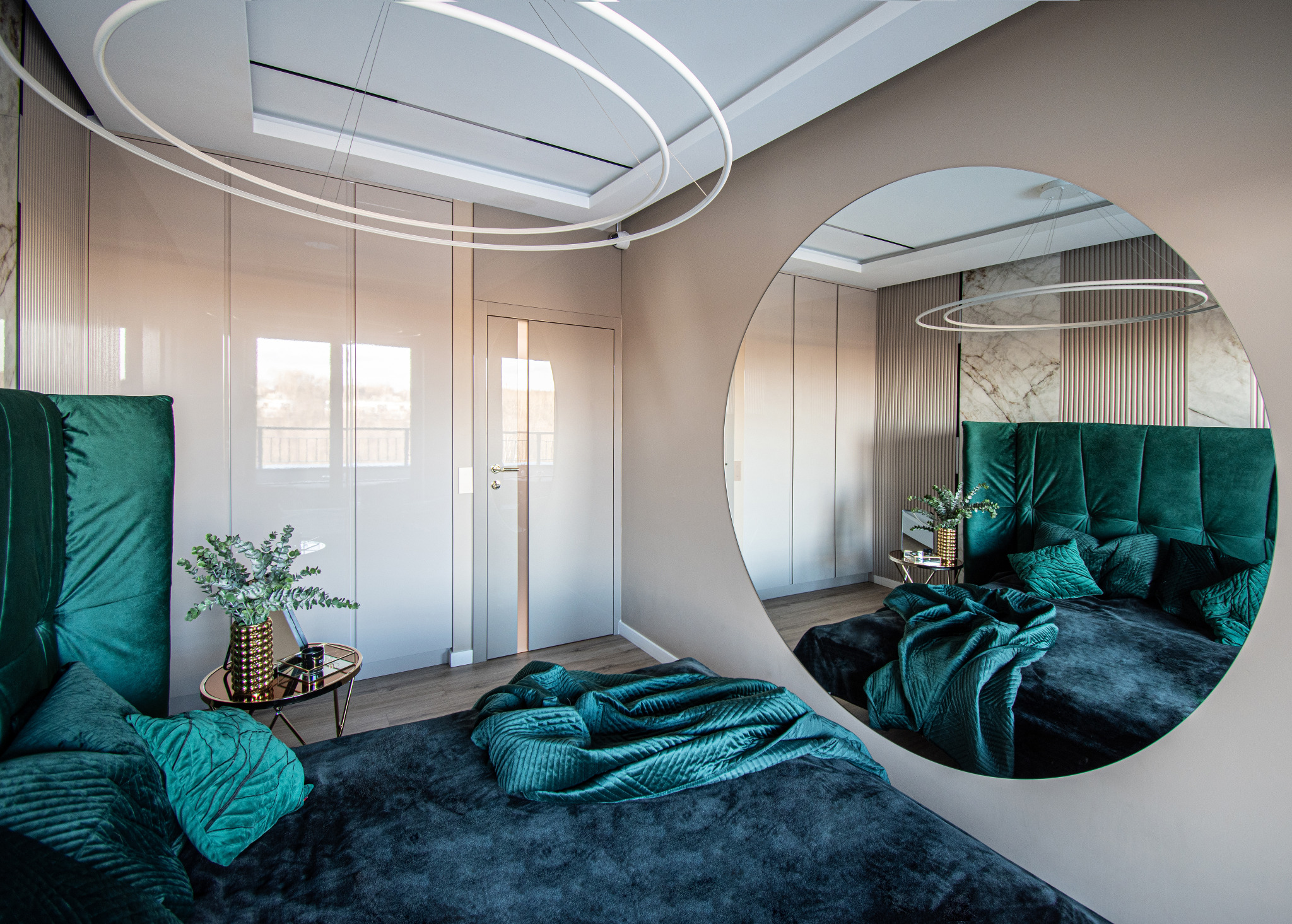 Elegancka sypialnia z łóżkiem z szmaragdowym zagłówkiem, okrągłym lustrem, stolikiem nocnym i nowoczesnym oświetleniem sufitowym.