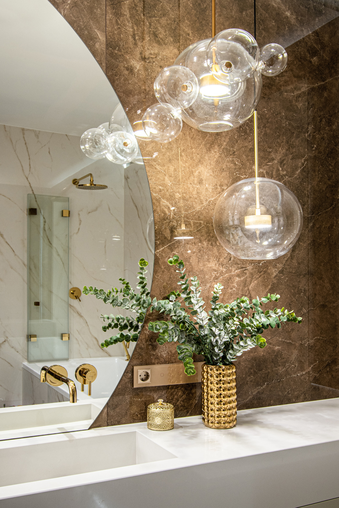 Luksusowa łazienka z marmurowymi ścianami, szklanymi lampami wiszącymi, złotą armaturą i wazonem z eukaliptusem na białym blacie umywalki.