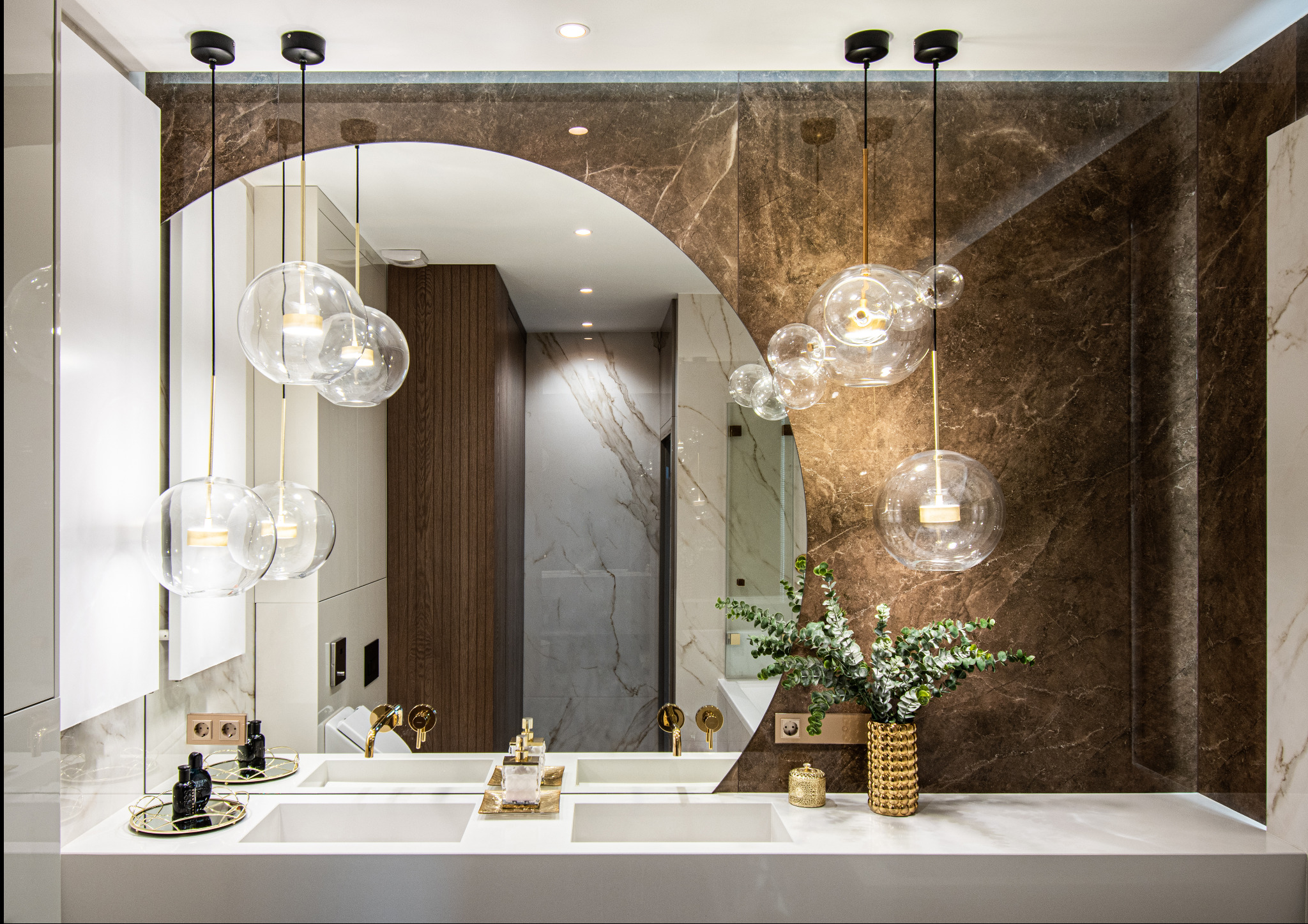 Elegancka łazienka z podwójną umywalką, okrągłym lustrem i marmurową ścianą w odcieniach brązu i bieli. Nad umywalką wiszą szklane lampy w kształcie kul, a na blacie stoją złote akcesoria i wazon...
