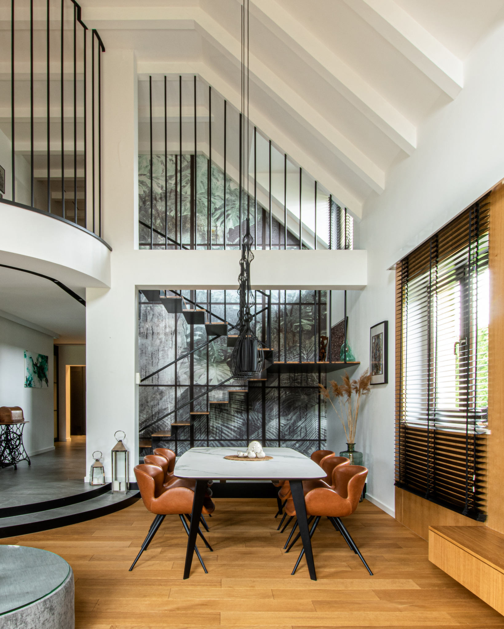 Dwupoziomowe wnętrze domu z antresolą, drewnianą podłogą i stołem jadalnym z krzesłami w kolorze koniakowym. W tle spiralne schody z motywem dżungli na ścianie i wisząca lampa z czarnych sznurków.