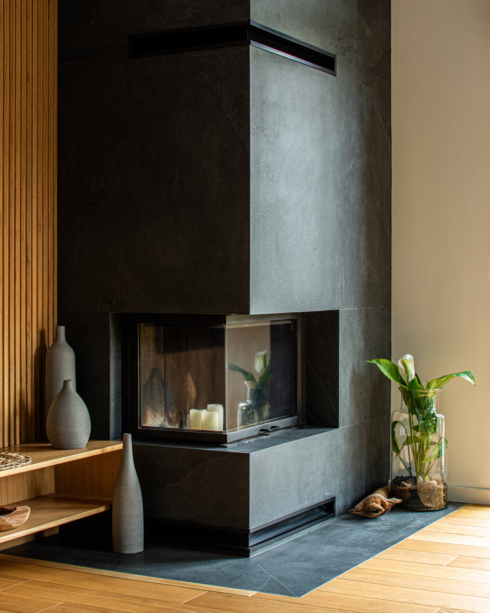 Nowoczesny kominek obłożony czarnym kamieniem, z paleniskiem zamkniętym szkłem, obok drewniane lamele i dekoracyjne wazony oraz szklany wazon z rośliną.