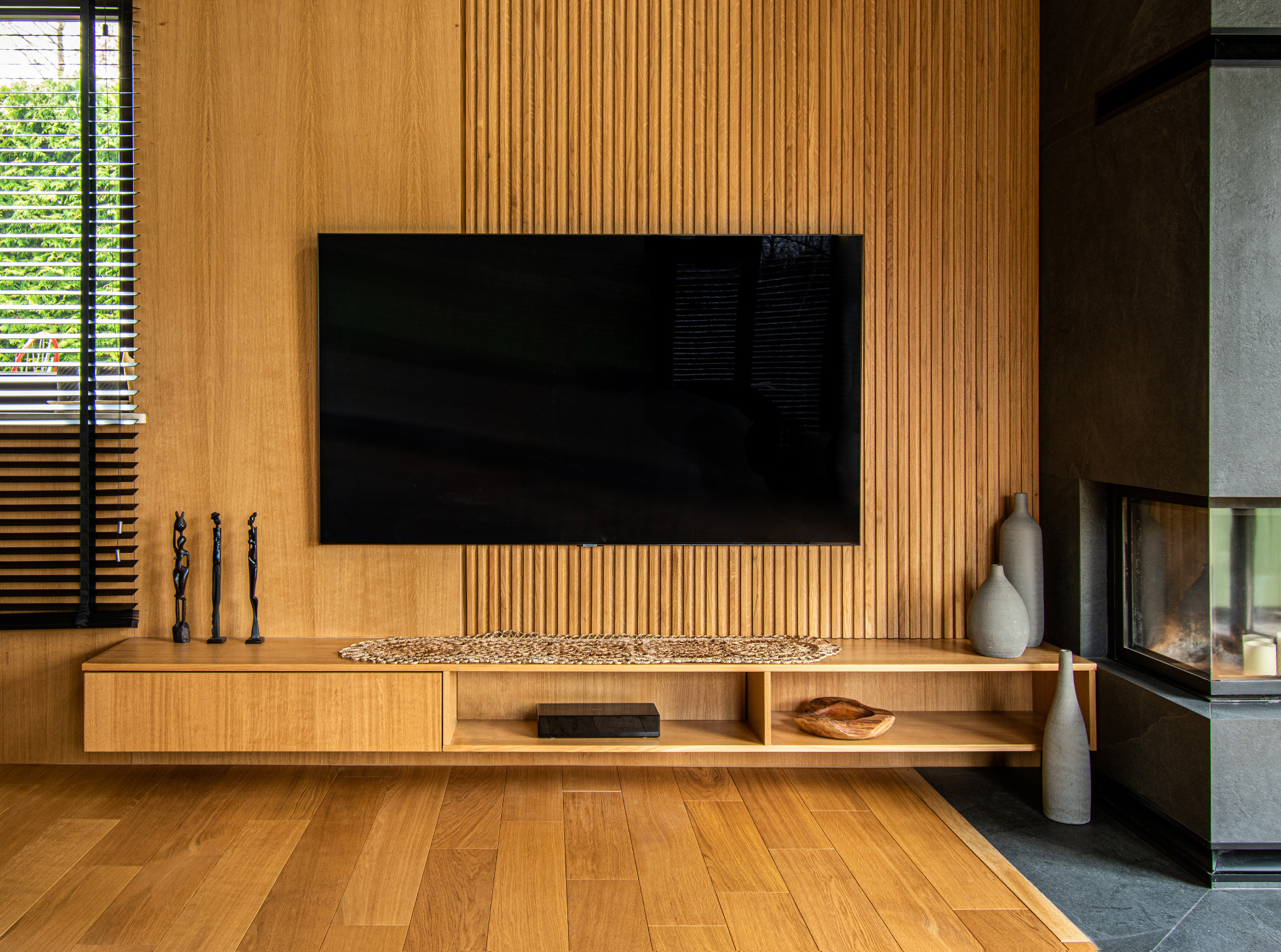 Nowoczesny salon z drewnianą ścianą, wiszącą szafką RTV, czarnym telewizorem, trzema afrykańskimi rzeźbami i kominkiem obłożonym ciemnym kamieniem.
