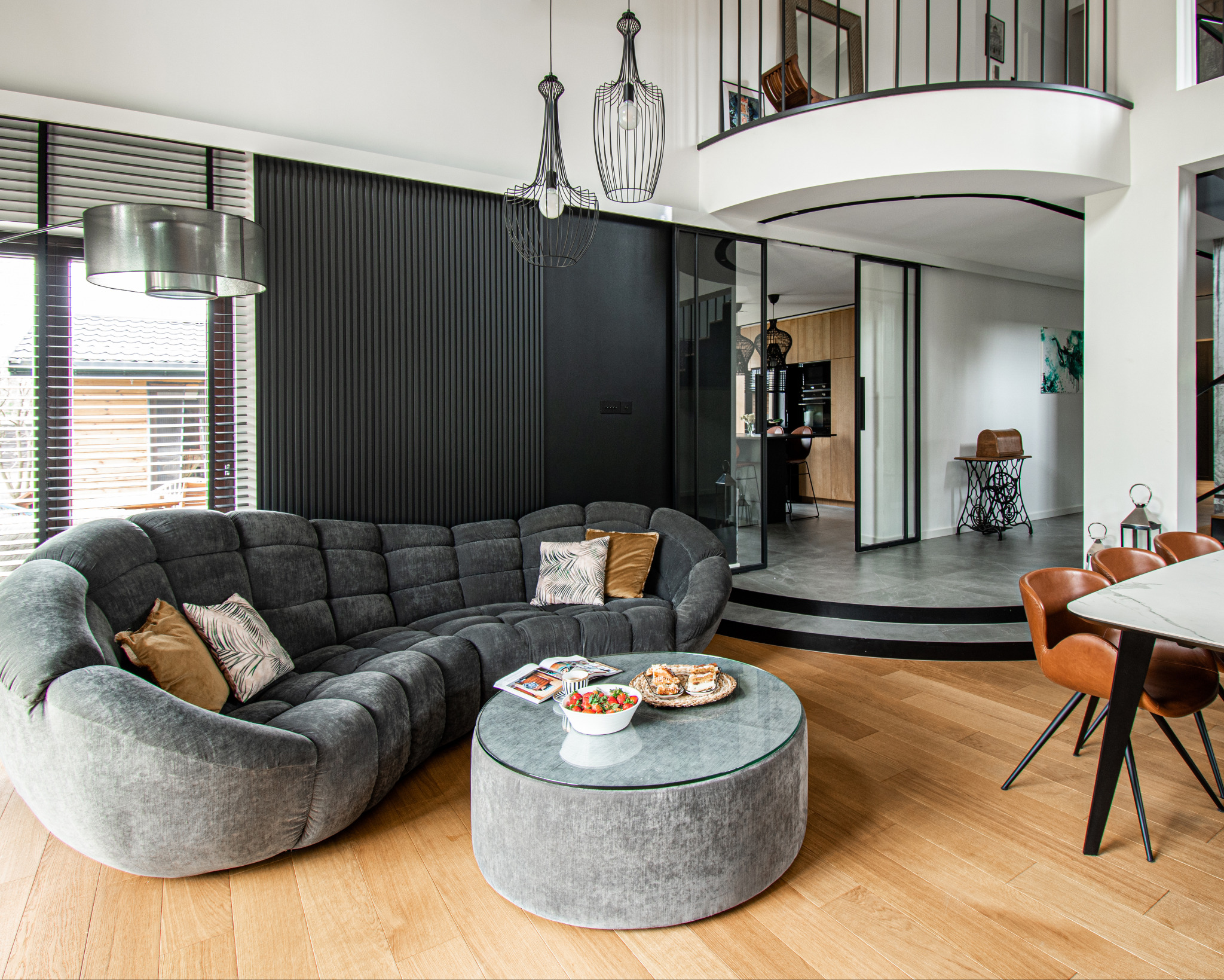 Nowoczesny salon z szarą sofą modułową, okrągłym stolikiem kawowym z przekąskami, czarną ścianą z pionowymi panelami oraz widokiem na kuchnię przez przesuwne szklane drzwi.
