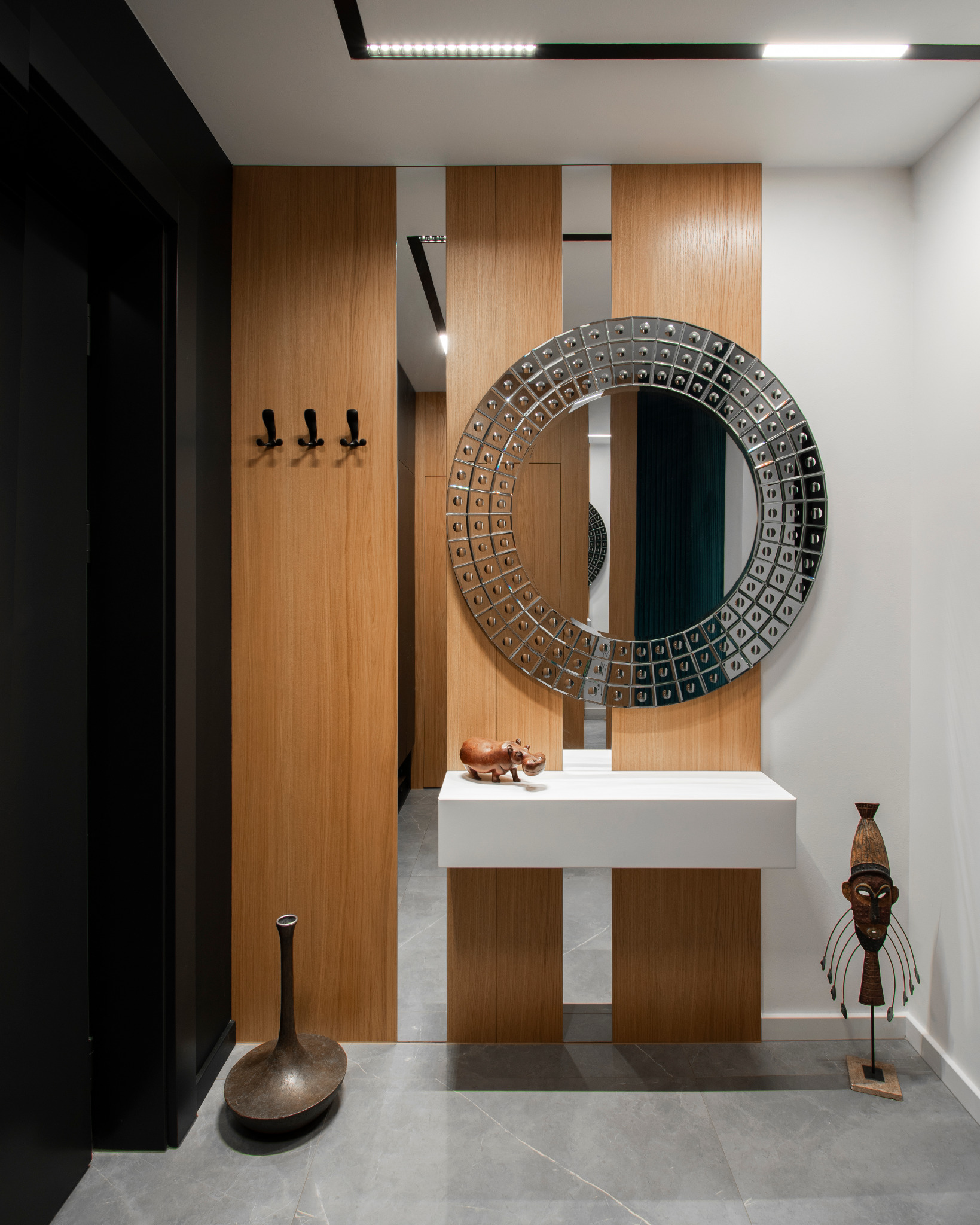Nowoczesny przedpokój z drewnianą boazerią i okrągłym lustrem dekoracyjnym, uzupełniony o afrykańską maskę i figurkę hipopotama.