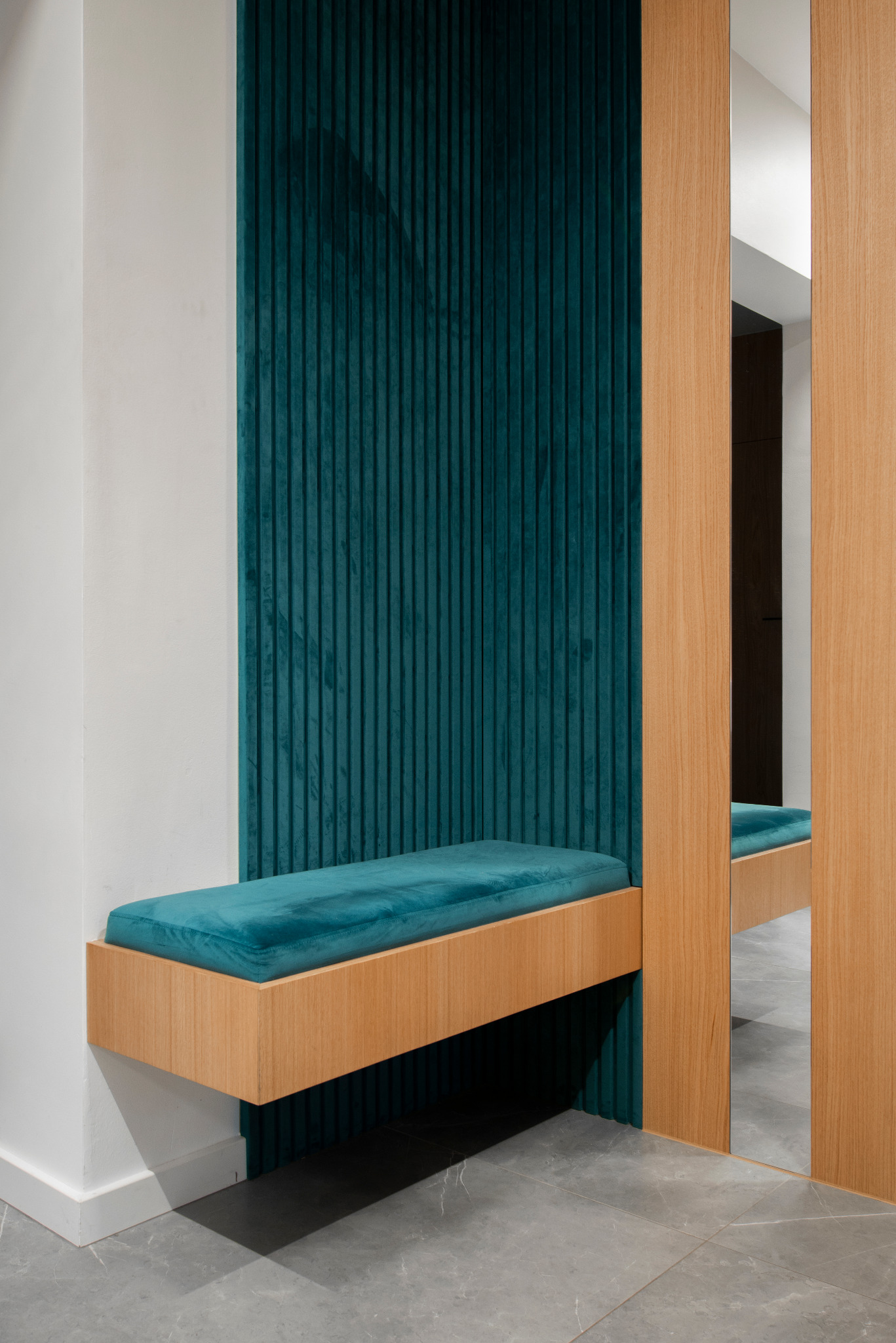 Nowoczesny przedpokój z siedziskiem obitym turkusowym welurem, drewnianą ramą i lustrem, na tle ściany z panelami w pionowe prążki w odcieniu butelkowej zieleni.
