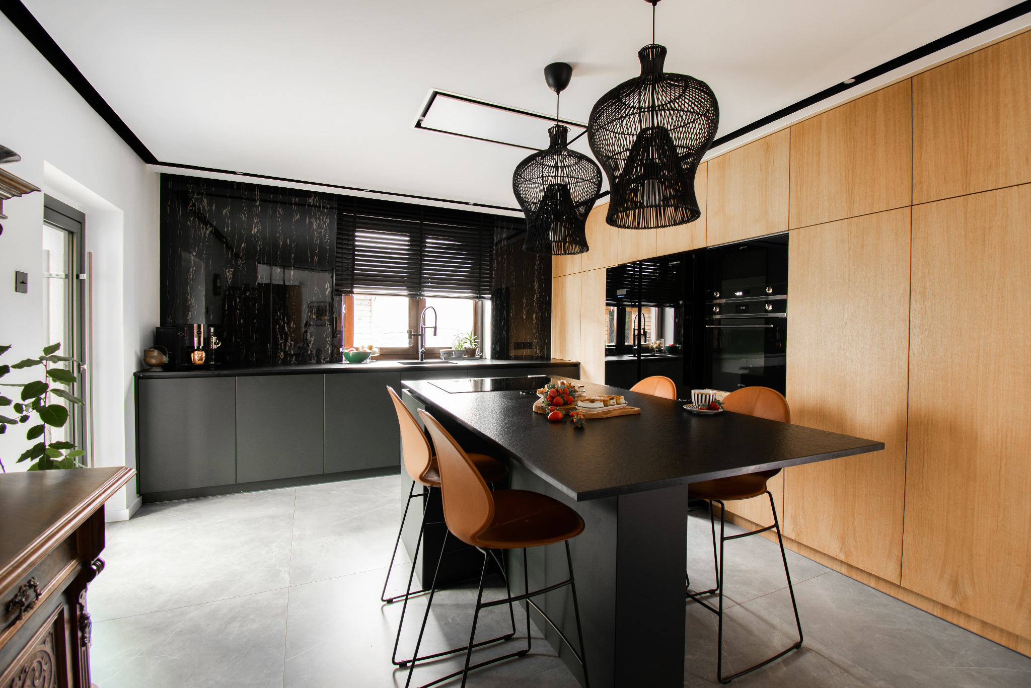 Nowoczesna kuchnia z czarnym marmurem, drewnianymi szafkami, wyspą z czarnym blatem i trzema plecionymi lampami wiszącymi nad nią, oraz z krzesłami barowymi w kolorze karmelowym.