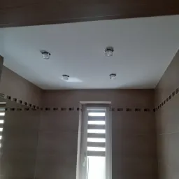 Wykończona łazienka z beżowymi płytkami, dekoracyjnym pasem mozaiki, czterema punktowymi lampami sufitowymi i oknem z żaluzjami poziomymi.