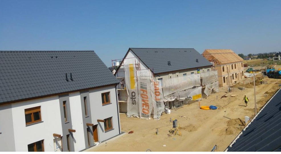 Osiedle w budowie: Domy jednorodzinne z szarą dachówką, jeden dom w trakcie ocieplania, drugi w stanie surowym z widoczną konstrukcją drewnianą, pracownicy w kamizelkach odblaskowych, sprzęt...