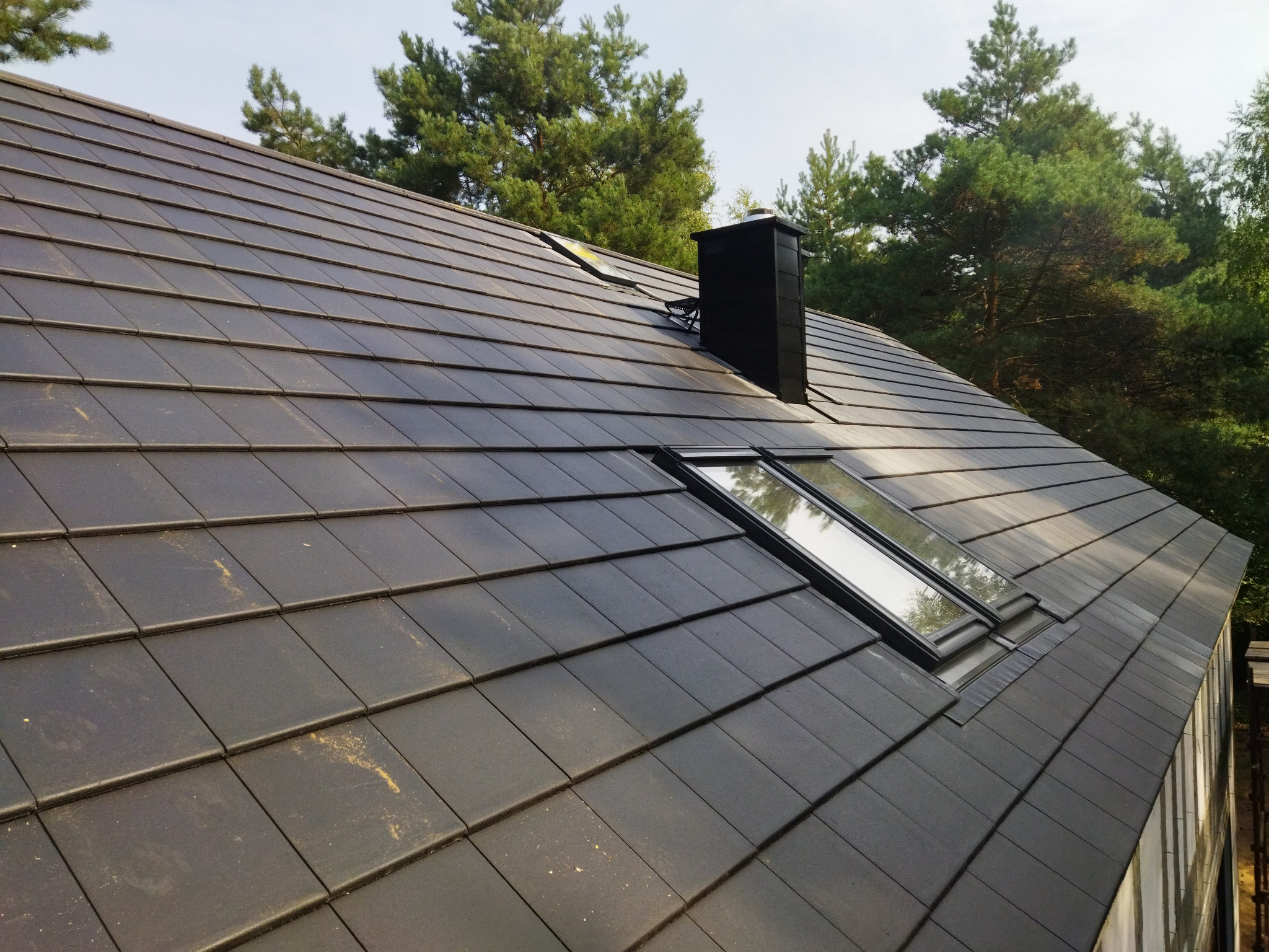 Ciemna dachówka ceramiczna ułożona na dachu z oknami dachowymi i kominem na tle zielonych drzew.