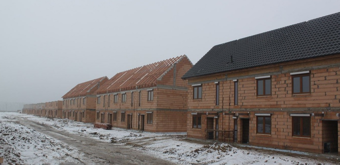 Szeregówka domów jednorodzinnych w trakcie budowy, z jednym domem z czarnym dachem i zamontowanymi oknami, pozostałe w stanie surowym z widoczną konstrukcją dachu, teren wokół pokryty śniegiem...