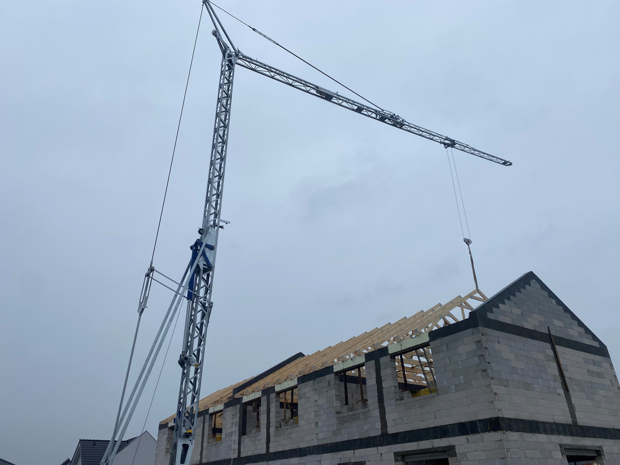 Wznoszenie domu z bloczków betonowych i drewnianej więźby dachowej przy użyciu wysokiego żurawia budowlanego w pochmurny dzień.