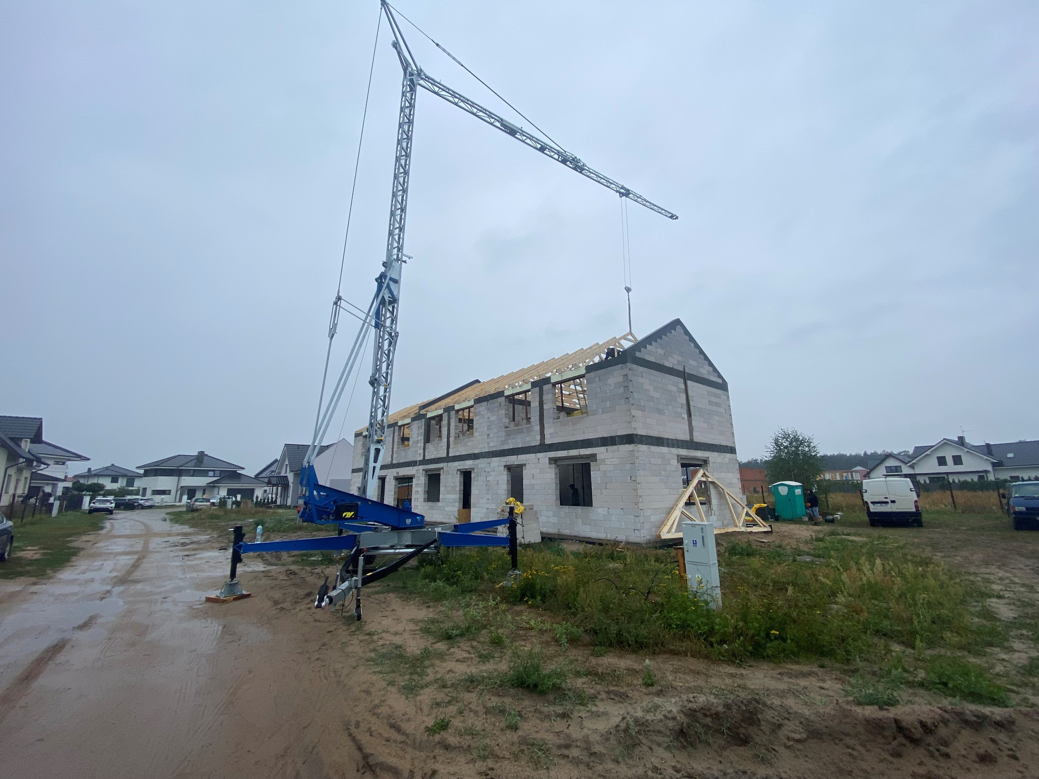 Budowa domu z bloczków: widok na dwukondygnacyjny budynek w stanie surowym otwartym, z widoczną konstrukcją dachu i dźwigiem budowlanym na pierwszym planie, z błotnistą drogą dojazdową.