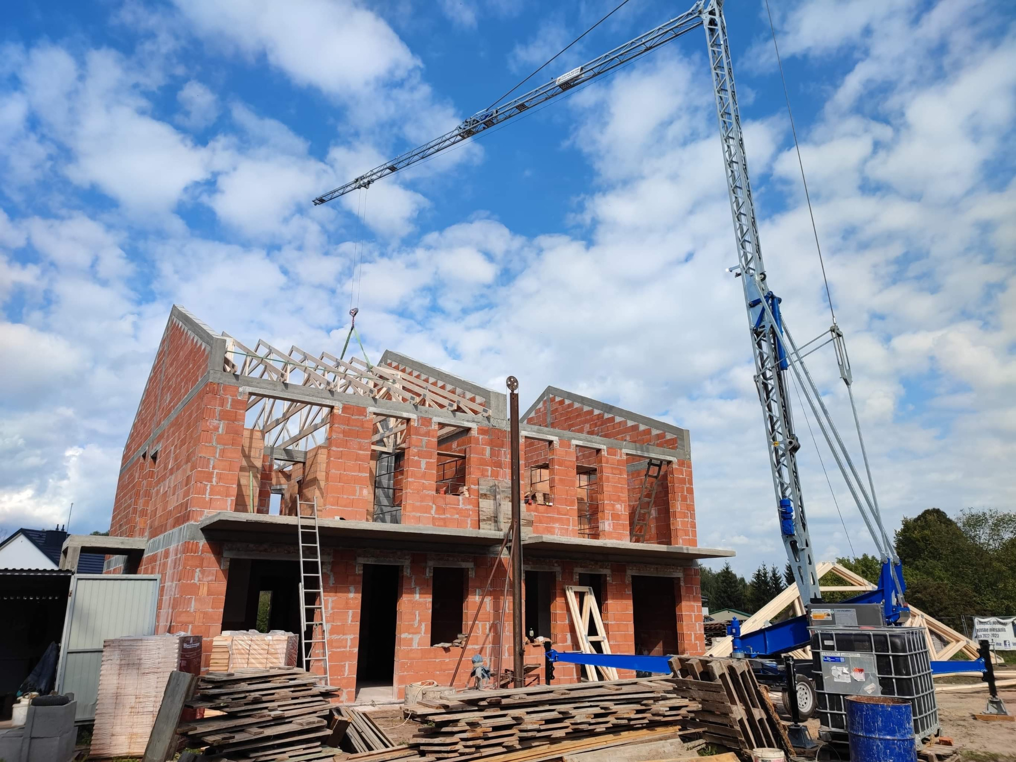 Budowa domu w stanie surowym otwartym z użyciem dźwigu, widoczne cegły, betonowe stropy, drewniana konstrukcja dachu oraz stos desek na pierwszym planie, błękitne niebo z chmurami w tle.