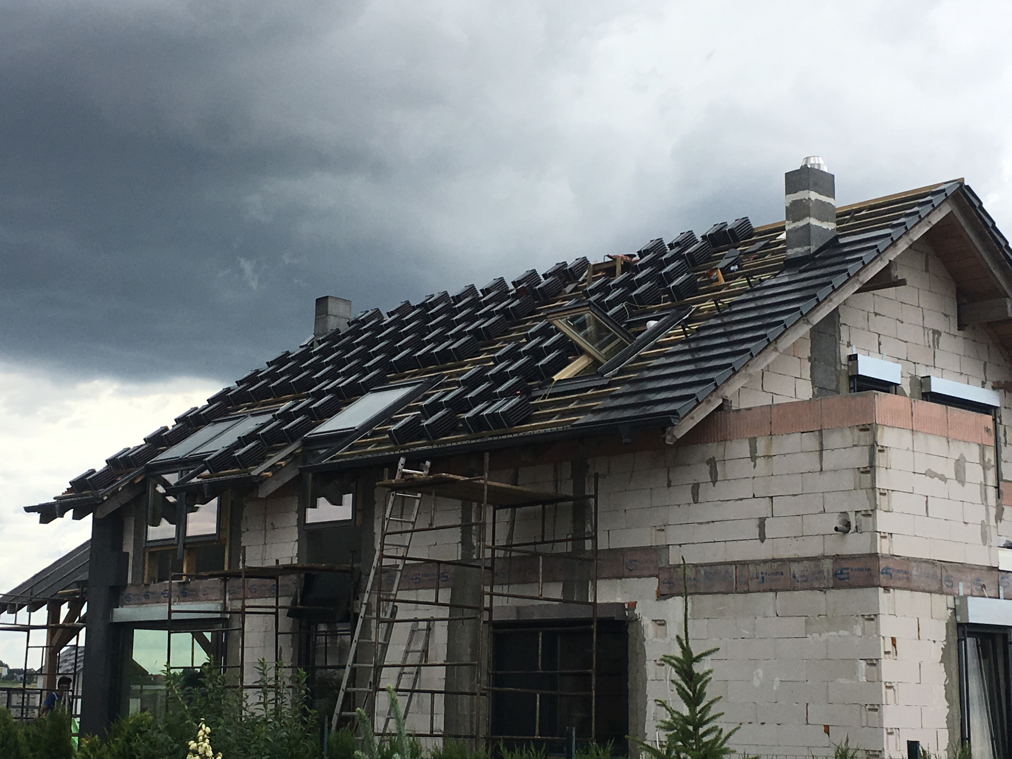 Dom w trakcie budowy z częściowo ułożonym ciemnoszarym dachem i oknami dachowymi, widoczne rusztowania i elementy konstrukcji, pochmurne niebo w tle.
