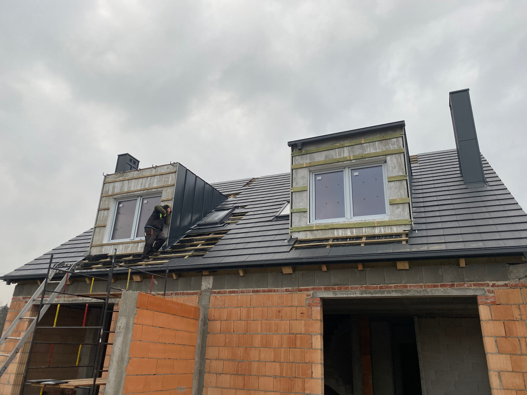 Budowa domu w trakcie krycia dachu dachówką ceramiczną, widoczny dekarz na dachu, okna dachowe i komin, surowy mur z cegły, drabina oparta o budynek, pochmurne niebo w tle.