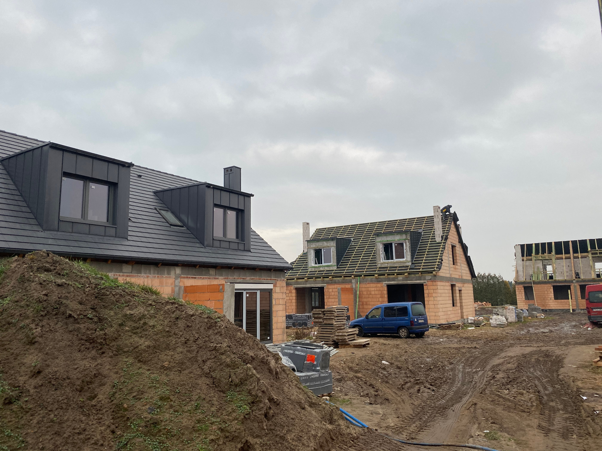 Budowa domów jednorodzinnych w trakcie realizacji: jeden dom z ciemnoszarą blachą na dachu i oknami dachowymi, drugi w trakcie krycia więźby dachowej, trzeci w stanie surowym. Na pierwszym planie...