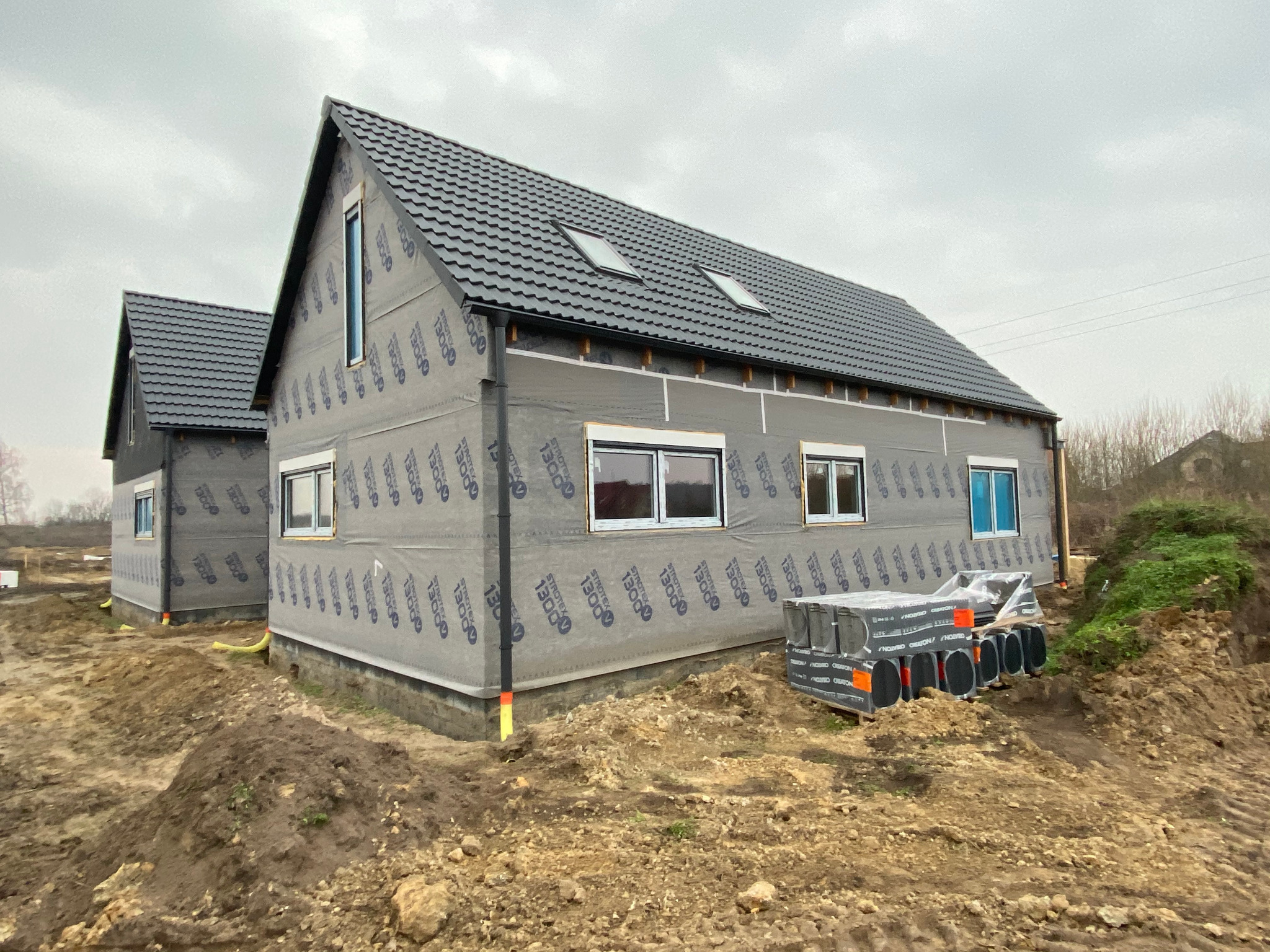 Dwa domy w trakcie budowy z czarną dachówką, zabezpieczone membraną. Na ziemi leżą zapakowane paczki materiałów izolacyjnych.