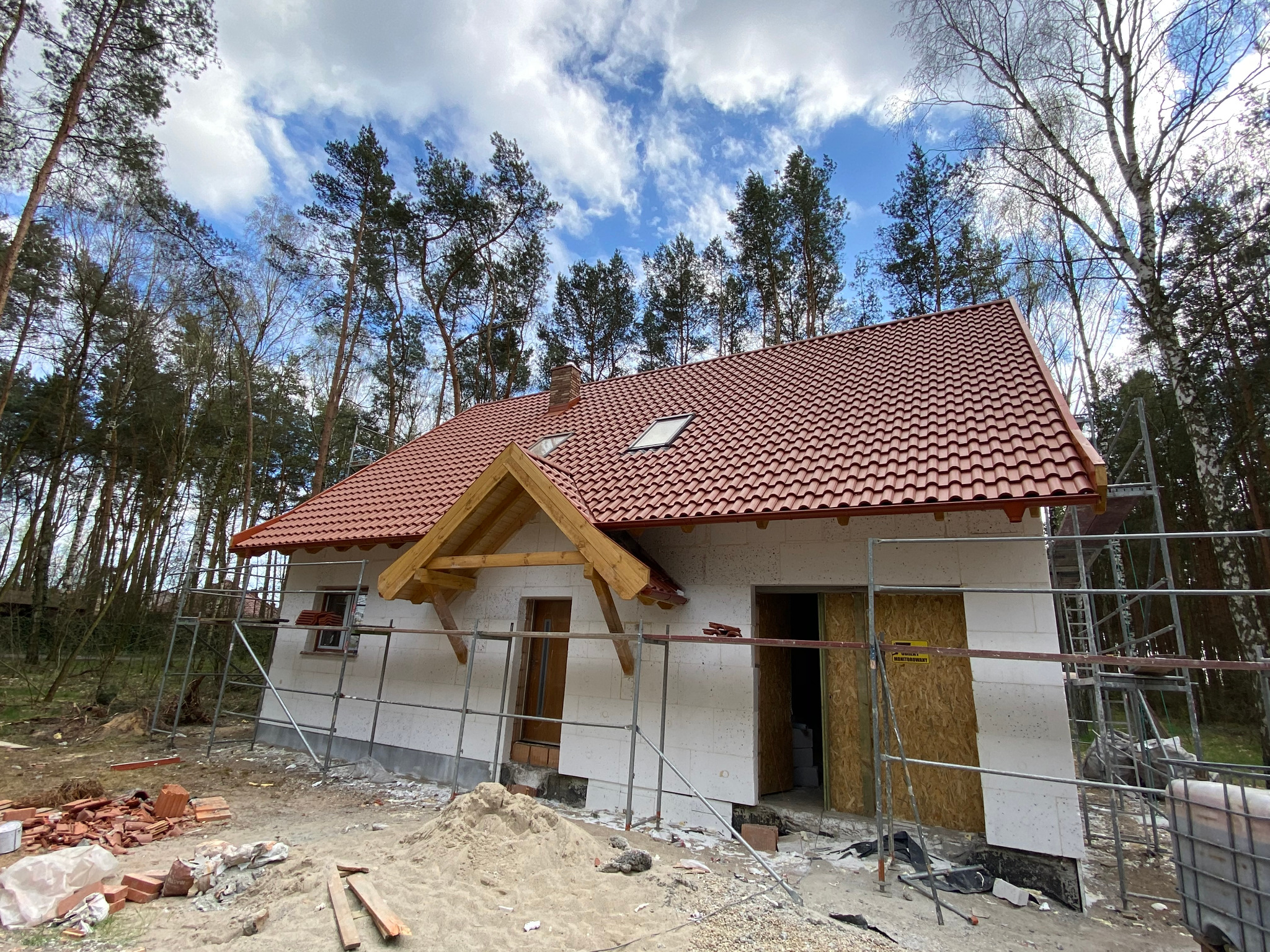 Niedokończony dom jednorodzinny w trakcie budowy, z czerwoną dachówką, widoczne rusztowania, drewniana konstrukcja zadaszenia nad wejściem oraz stosy cegieł na ziemi.