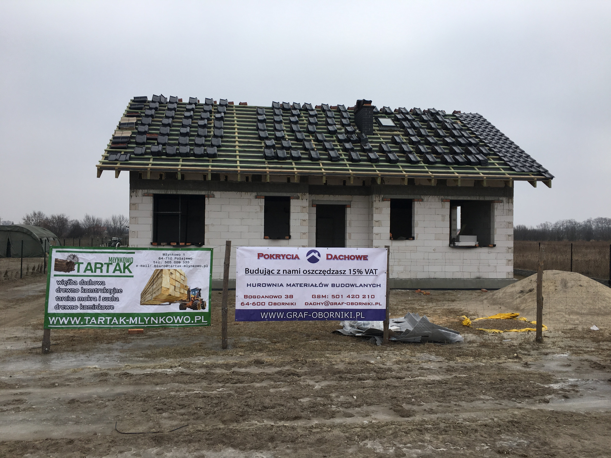 Dom w trakcie krycia dachu dachówką ceramiczną, widoczna konstrukcja więźby dachowej i częściowo ułożona dachówka, na pierwszym planie reklamy firm budowlanych i tartaku.