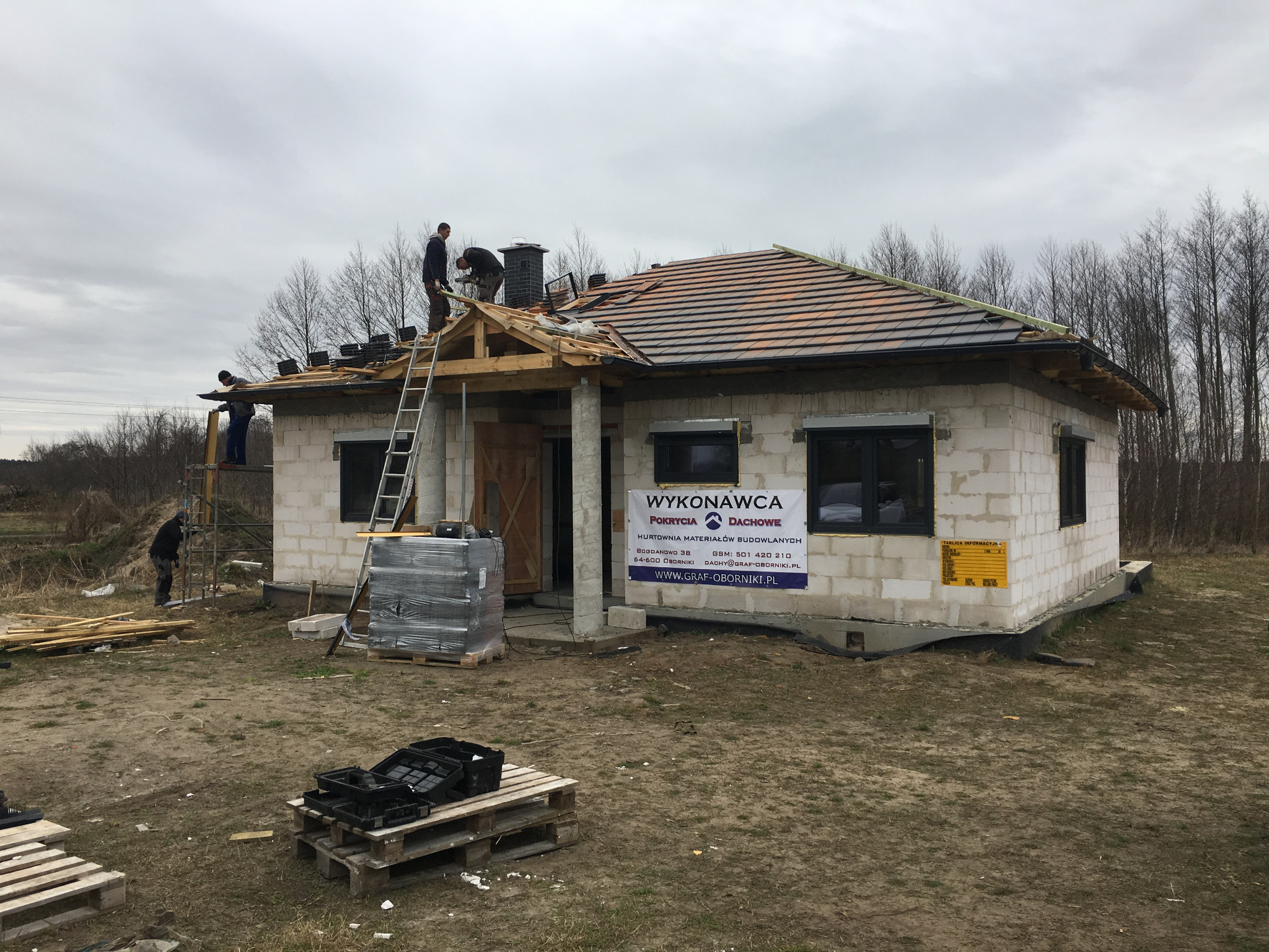 Ekipa budowlana układa dachówkę na nowym domu z bloczków, widoczna więźba dachowa i częściowo pokryty dach, na elewacji baner reklamowy firmy dekarskiej.