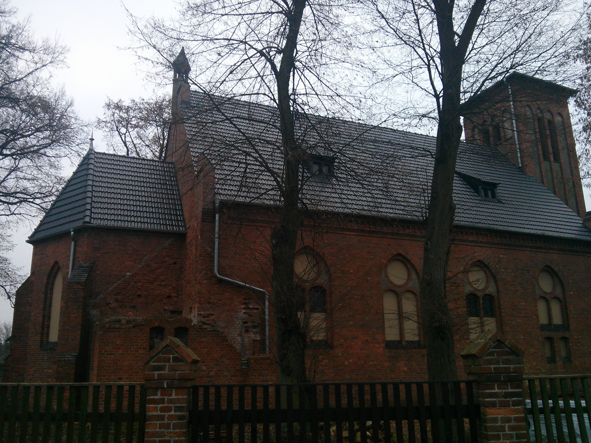 Ceglany kościół z nową dachówką, widoczny zza drewnianego płotu i drzew bez liści, w pochmurny dzień.