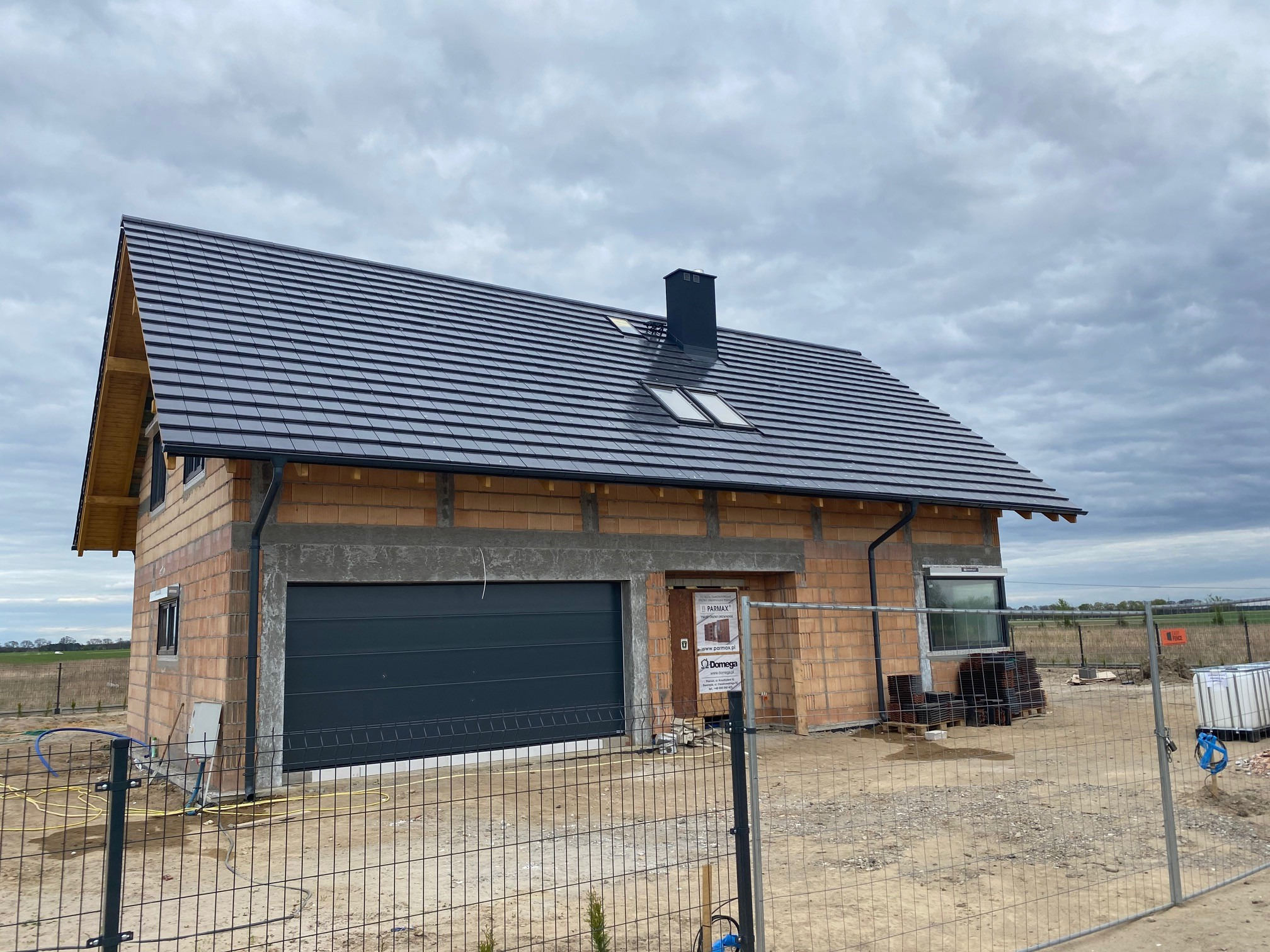 Niedokończony dom jednorodzinny w trakcie budowy z ciemnoszarą dachówką, widoczne okna dachowe i komin, ściany z pustaków ceramicznych, brama garażowa, teren ogrodzony siatką.