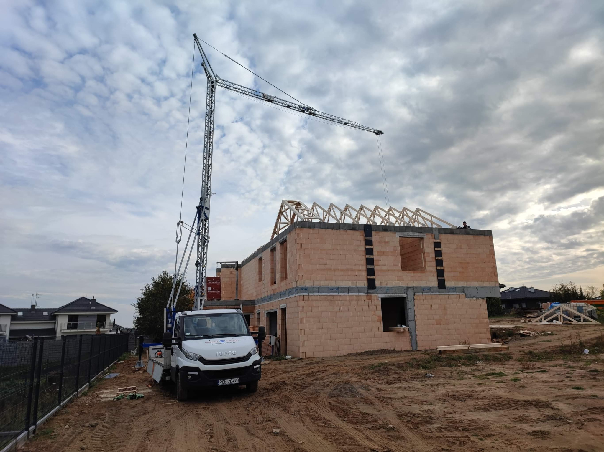Wznoszenie budynku z bloczków ceramicznych z wykorzystaniem żurawia samochodowego; widoczna konstrukcja więźby dachowej i przygotowane elementy na ziemi.
