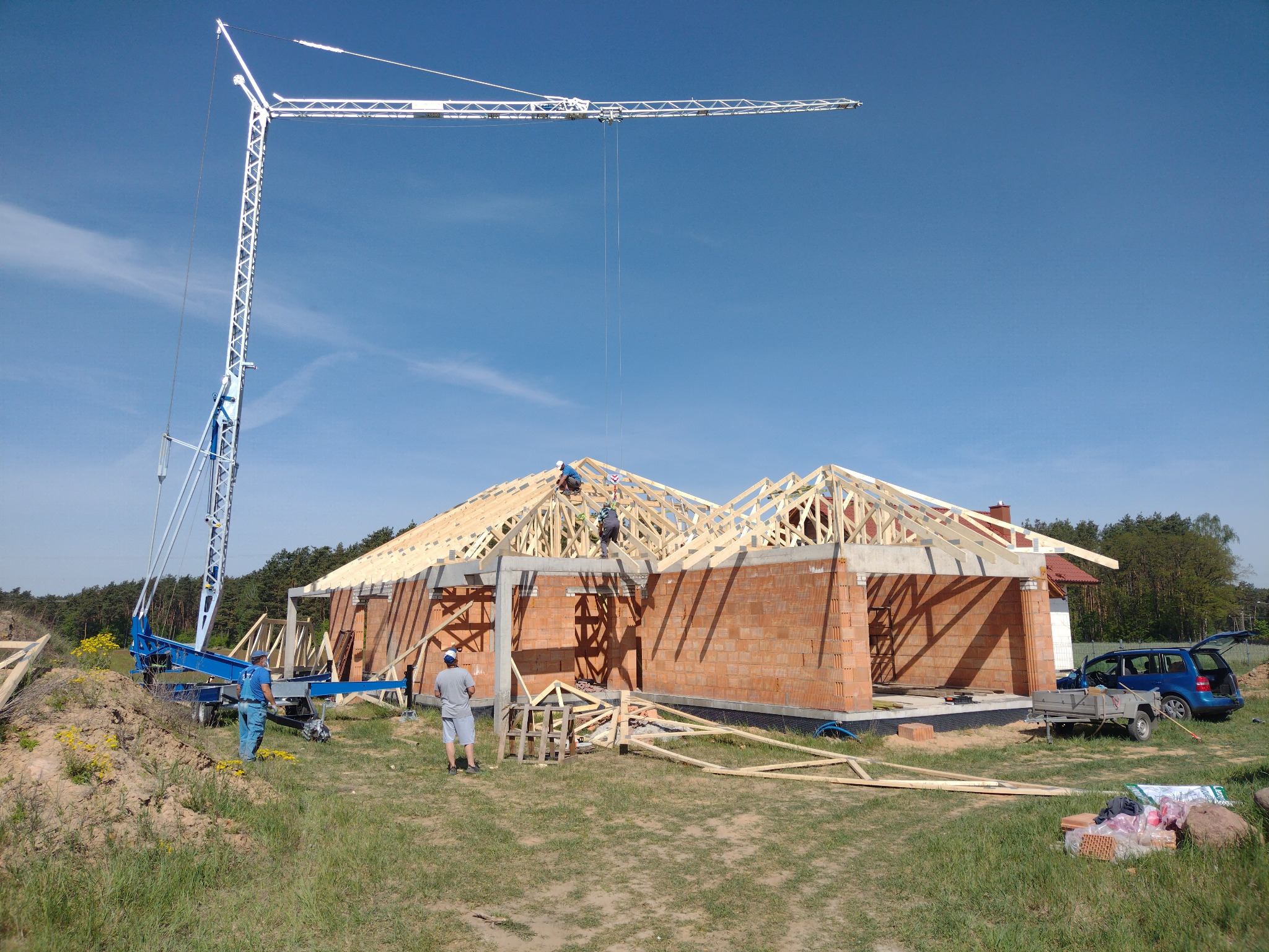Budowa domu jednorodzinnego w trakcie montażu drewnianej konstrukcji dachu, widoczny dźwig budowlany, pracownicy na dachu, oraz częściowo wymurowane ściany z czerwonej cegły.