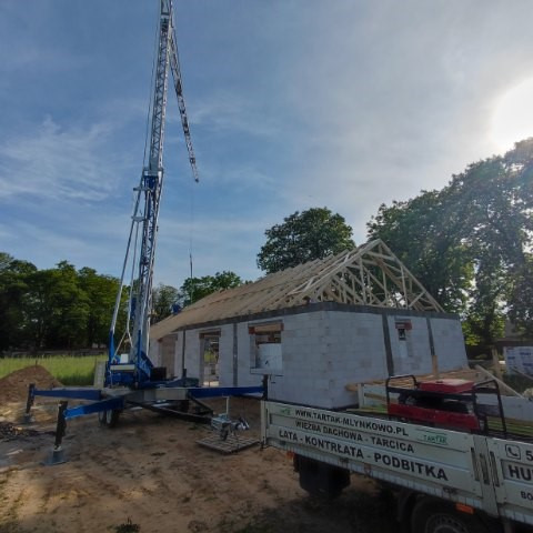 Budowa domu jednorodzinnego w trakcie wznoszenia więźby dachowej, widoczny żuraw budowlany i ciężarówka z materiałami, słoneczny dzień.