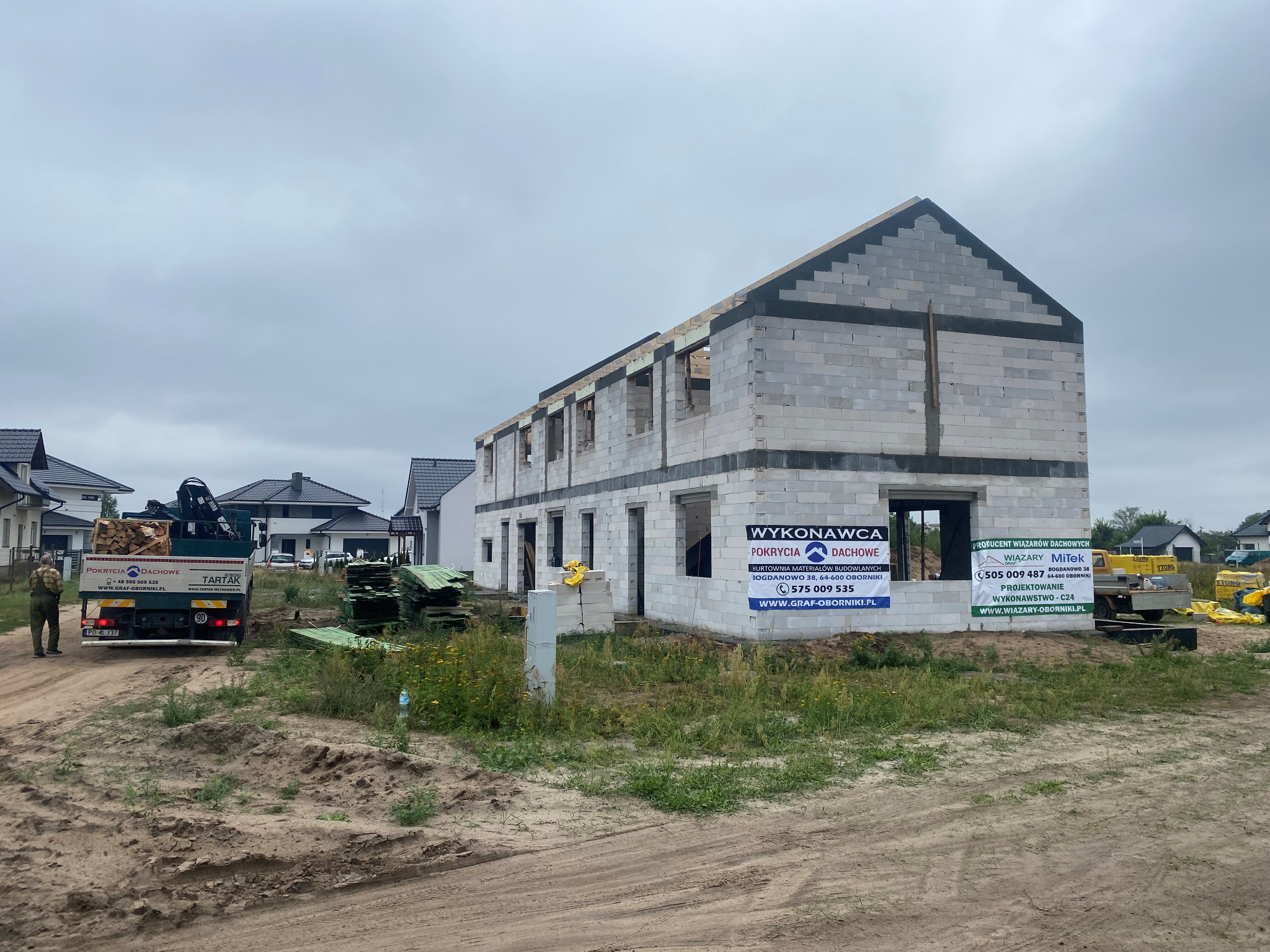 Surowy stan budynku w trakcie budowy, ściany z betonu komórkowego, widoczna więźba dachowa, obok zaparkowany samochód dostawczy z drewnem, w tle domy jednorodzinne.