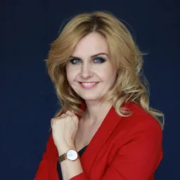 Portret uśmiechniętej kobiety z blond włosami, w czerwonej marynarce i z zegarkiem na ręce, na ciemnoniebieskim tle.