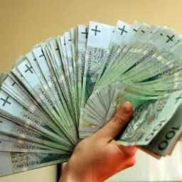 Dłoń trzymająca wachlarz banknotów 100 złotowych na rozmytym, beżowym tle.