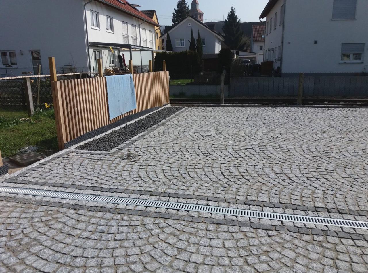 Szara kostka granitowa ułożona w łukowaty wzór, oddzielona liniowym odpływem wody, z drewnianym ogrodzeniem w tle i niebieskim ręcznikiem na nim.