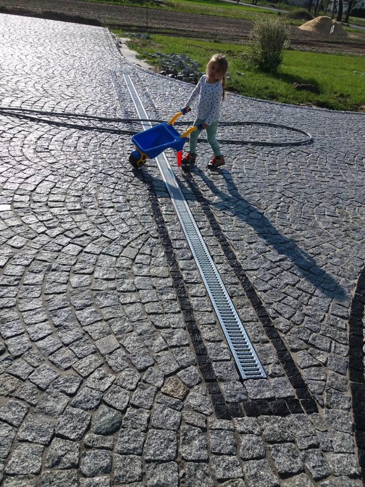 Ułożona kostka granitowa w łukowaty wzór z odpływem liniowym; dziewczynka bawi się wózeczkiem.