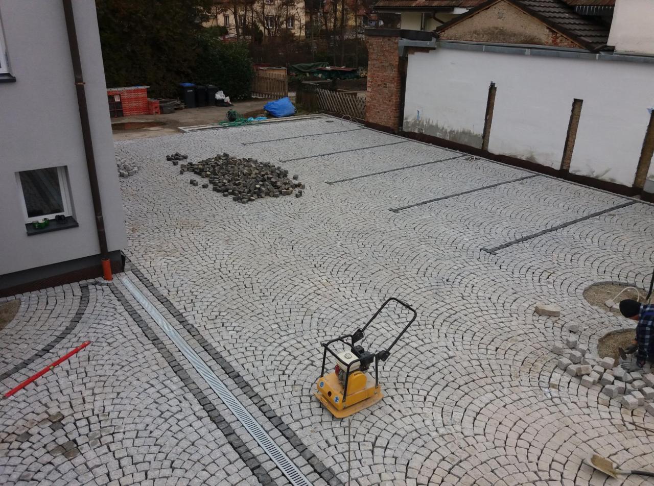 Układanie szarej kostki na podjeździe z widocznymi liniami drenażowymi i stertą kostki do ułożenia. Żółta zagęszczarka w centrum kadru.