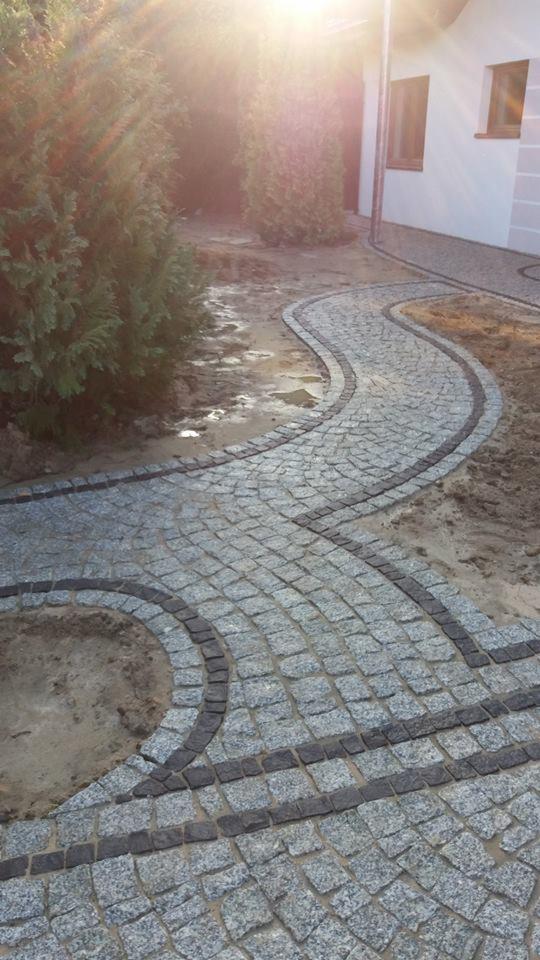 Kręta ścieżka z jasnoszarej kostki granitowej z ciemnoszarym obramowaniem, prowadząca przez ogród w kierunku białego budynku. Widoczne ślady po deszczu i przygotowane miejsce na nasadzenie roślin.