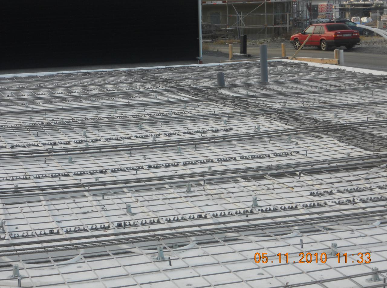 Siatka zbrojeniowa ułożona na białych płytach izolacyjnych, wystające rury kanalizacyjne, widoczny samochód osobowy w tle.