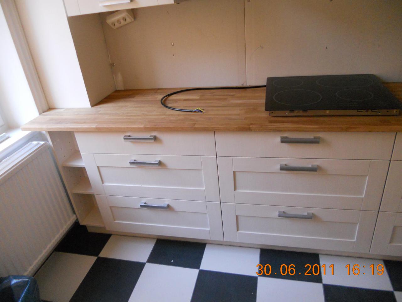 Kuchnia z białymi szafkami z szufladami, drewnianym blatem, czarną płytą grzewczą i czarno-białą podłogą w szachownicę, widok z przodu.