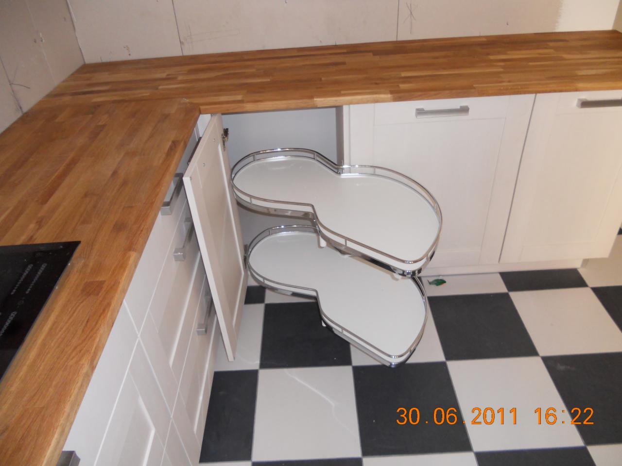 Wysuwany, dwupoziomowy system półek narożnych w otwartej szafce kuchennej z białymi frontami, drewniany blat, czarno-biała podłoga w szachownicę.