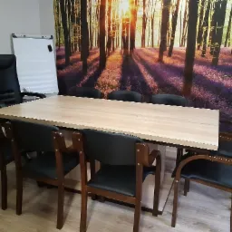 Sala konferencyjna z dużym stołem, krzesłami z czarnej skóry i fototapetą przedstawiającą las w tle.