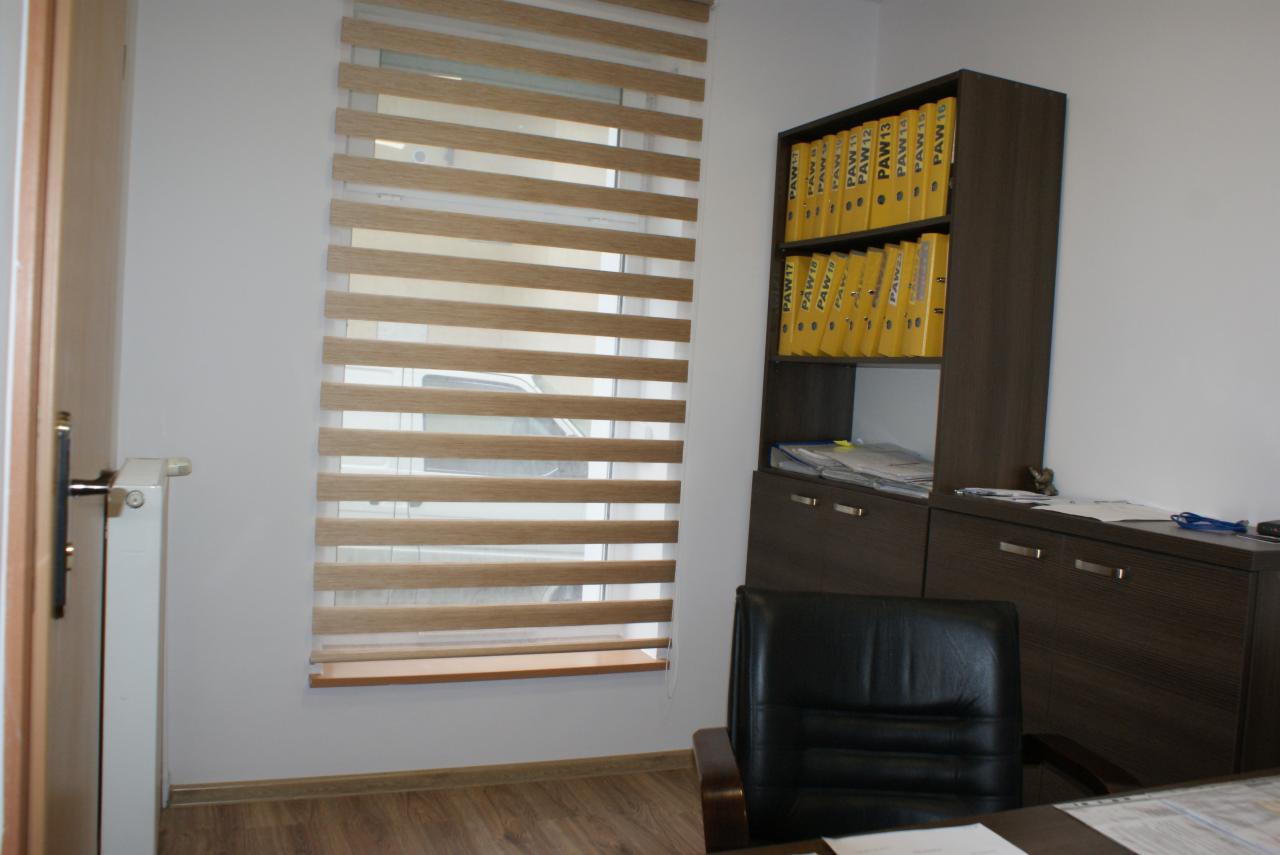 Wnętrze biura z biurkiem, krzesłem, regałem z segregatorami oznaczonymi PAW i żaluzjami poziomymi w oknie.