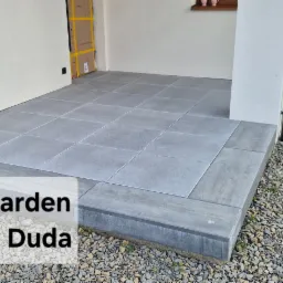 Wykończony taras z szarych płyt na podbudowie z kostki, obok ścieżka z kamyków i trawnik. Widok z boku, w tle fragment budynku.