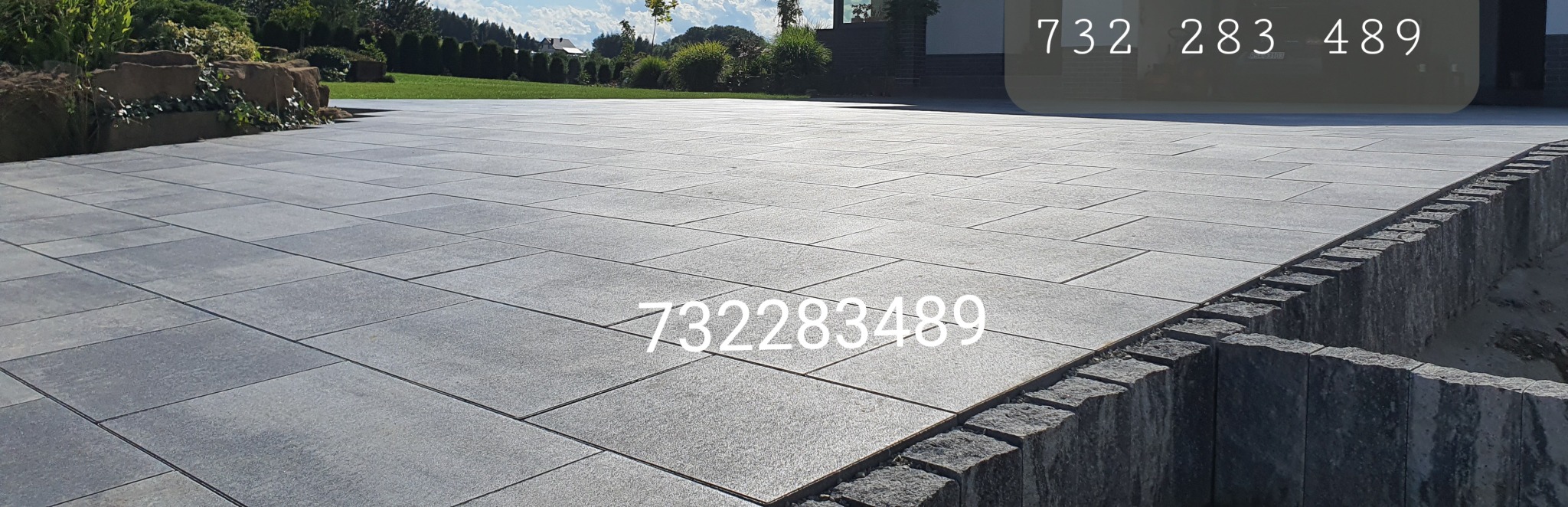 Szara, prostokątna nawierzchnia z kostki, z widocznym obrzeżem z kamiennych bloków i trawnikiem w tle.