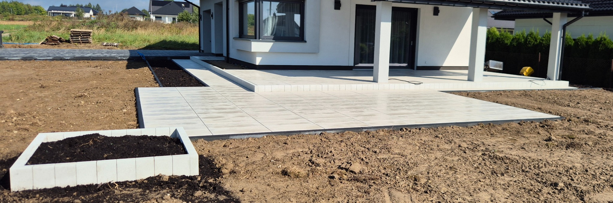 Jasna, prostokątna płyta tarasowa przylegająca do białego domu, obok rabata z ziemią. Widok z poziomu gruntu na ogród w trakcie realizacji.