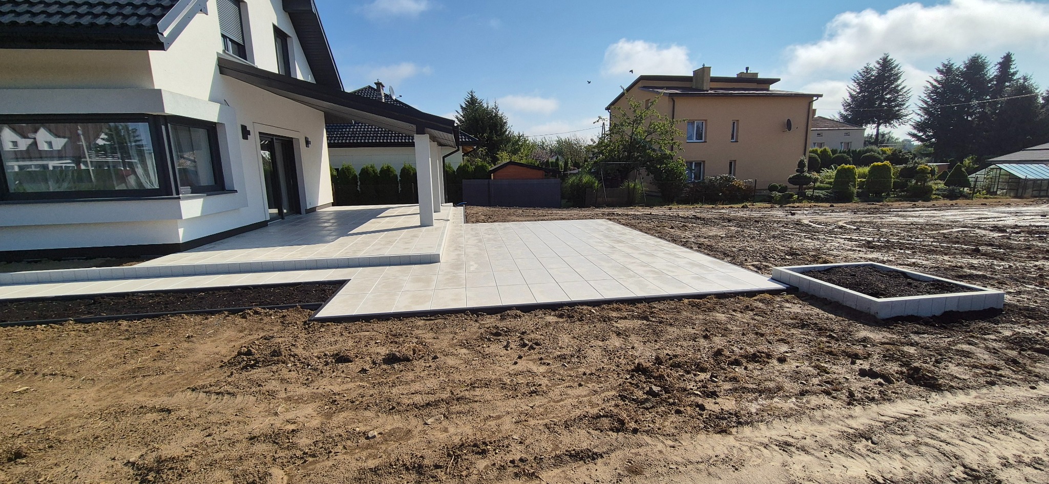 Jasna, kwadratowa kostka tarasowa ułożona przy nowoczesnym domu z białymi kolumnami, obok rabata. Grunt przygotowany pod dalsze prace ogrodowe.