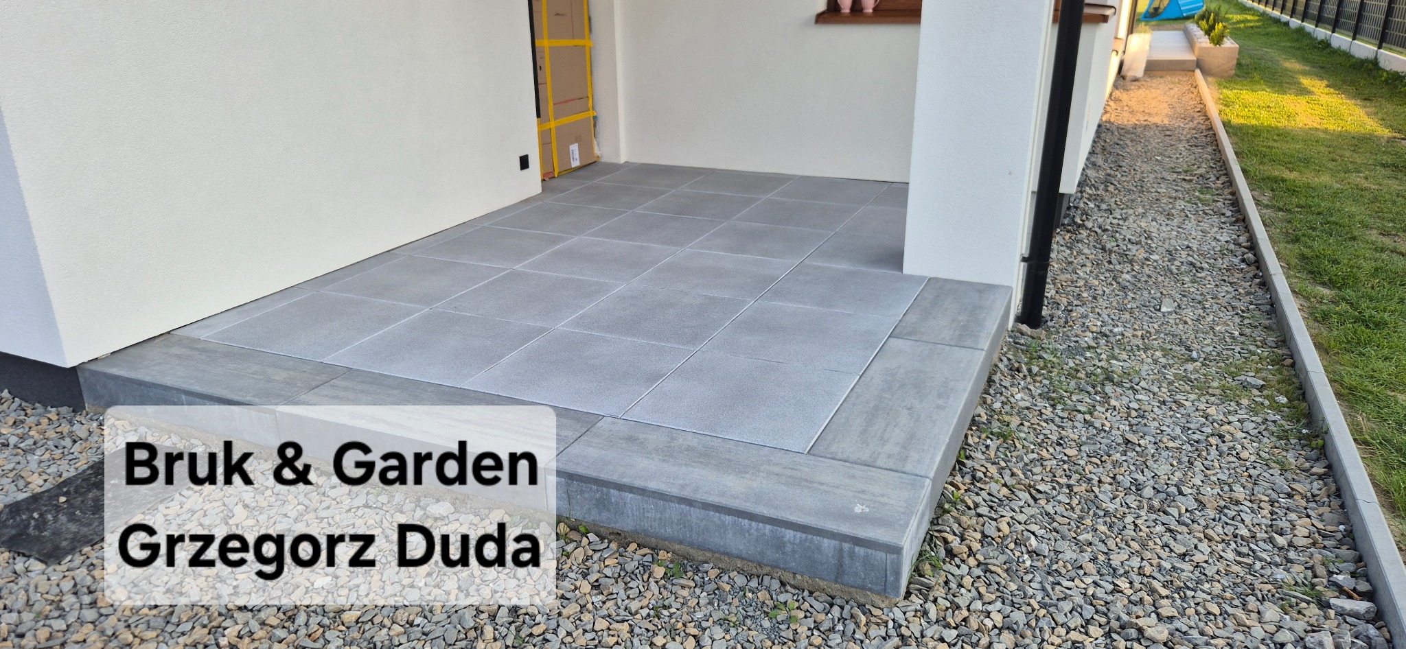Wykończony taras z szarych płyt na podbudowie z kostki, obok ścieżka z kamyków i trawnik. Widok z boku, w tle fragment budynku.