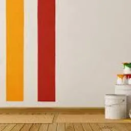 Świeżo pomalowana ściana z trzema pionowymi pasami w kolorach zielonym, żółtym i czerwonym. Obok ściany wałek do malowania i kilka białych wiader z farbami.